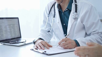 Doctor and a patient. The physician, wearing a white medical coat over a green shirt, is filling out a medical record form during a consultation in the clinic. Medical service