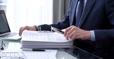 Businessman using a laptop computer and magnifying glass while analyzing financial documents at desk in modern office. Audit and taxes in business