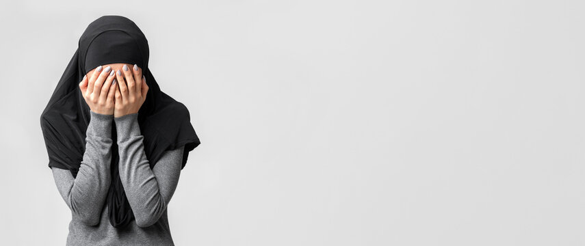 Islamophobia and muslim women discrimination concept. Woman in black hijab covering face with hands, posing over gray studio background
