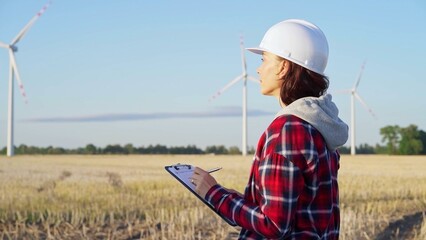 Female engineer wearing a white protective helmet is taking notes with a clipboard in a field with wind turbines, as the sun sets. Clean energy concept