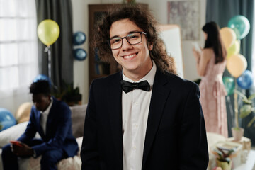Portrait of curly-haired young man smiling at camera while wearing suit and bowtie at social gathering or party event in indoor setting