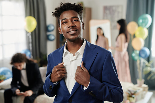 Portrait of smiling young man standing indoors adjusting his suit jacket at a party with balloons and people in the background