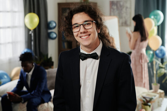 Portrait of curly-haired young man smiling at camera while wearing suit and bowtie at social gathering or party event in indoor setting