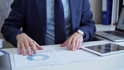 Businessman with blue suit is analyzing financial data using charts and graphs on paper and a tablet. Business and audit