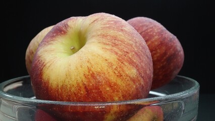 Close-up showcases a crisp bright red fresh apple nestled comfortably in a glass bowl. The black background creates a dramatic stage, highlighting the apple's flawless and smooth skin. Comestible.