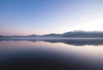 Serene Lake at Sunrise: A calm lake reflecting the soft hues of dawn, surrounded by misty mountains.