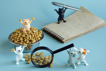 Toy kittens next to a bowl filled with dry cat food, a magnifying glass, a notebook and a pen. Selection of the best and tastiest pet food. Analysis of nutritional properties of animal food