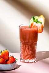 Non-alcoholic mocktail drink with strawberry, banana, syrup, peach juice, lemon and ice in glass. Beige pink background, hard light