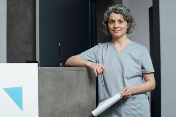 Portrait of medical professional standing in clinic, holding pen and paper in grey scrubs, smiling slightly, showing confidence
