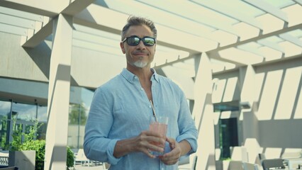 Satisfied smiling man in sunglasses holding, drinking refreshing pink cocktail on sunny terrace. Relaxed older male standing outdoor with happy smile.