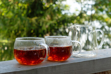 Two glass mugs with black hot tea and teapot on wooden table against forest background