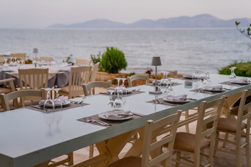 Nice greek style restaurant dining tables next to the Saronic islands in Greece and mountains in the background. No people and ready tables in the evening of the coast restaurant