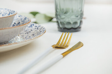 A view of a part of a set of white plates with a blue pattern on the edges stand on top of each other, a knife and fork are on the sides, a blue glass and a green sprig of sprouting on a white