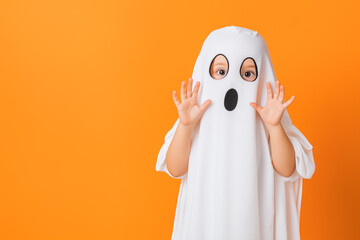 A young child wearing a ghost costume with a white sheet and spooky face cutouts, playfully posing against a vibrant orange background