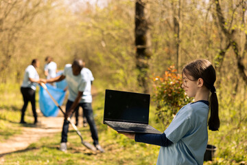 Small girl using isolated display on laptop during litter cleanup session in the woods, taking part in an environmental conservation activity. Little activist protecting the ecosystem and wildlife.