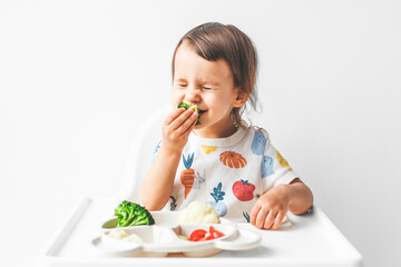 a happy child 1.5 years old sits in a high chair on a white background with a plate of vegetables, eats broccoli and grimaces, closing his eyes, concept of baby food and complementary feeding