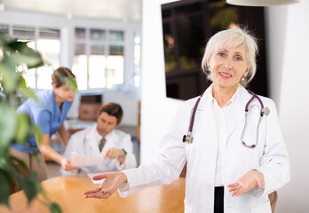 Fototapeta premium Smiling friendly elderly female doctor in white coat making welcome gesture, politely inviting patients in medical office ..