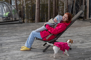 Caucasian woman with her son in an ergo backpack sitting in a wooden deck chair. 