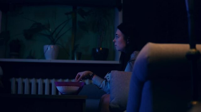 Woman is sitting in the living room on cozy sofa and watching TV eating nachos in the evening. Slow motion view of young adult woman spending time alone in front of television and having crisps
