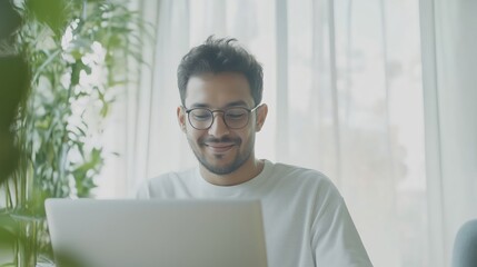 Young man with glasses smiling while working on a laptop from home, enjoying remote work in a comfortable and relaxed environment.