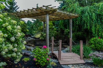 Bridge leads to pergola surrounded by lush foliage and flowers for backyard landscape design