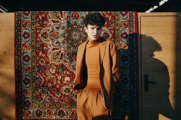 A young person in a stylish brown suit standing against a colorful patterned rug background, showcasing a blend of contemporary and traditional design elements