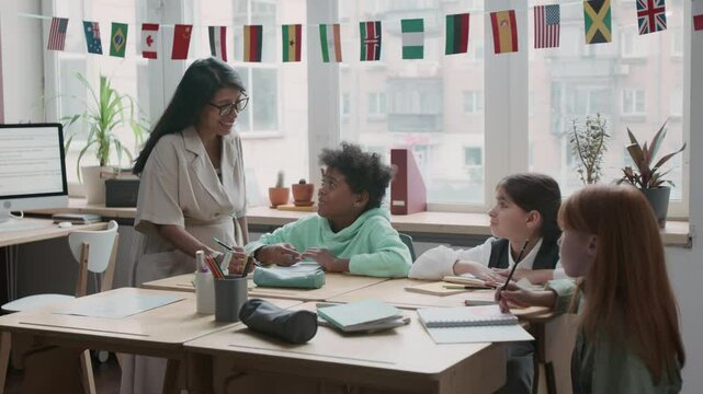 Medium shot of smiling Hispanic female teacher talking to 10-year-old African American schoolboy having difficulty with test at foreign language lesson, looking at his notebook and explaining task