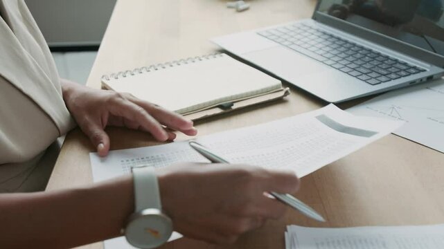 Close-up shot of hands of anonymous female teacher sitting at desk with laptop computer and checking students exam sheets while working alone in office