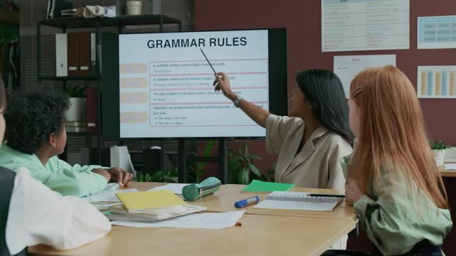 Medium shot of cheerful female Hispanic teacher explaining new English grammar rules to group of multiethnic schoolchildren, pointing at slide on multimedia screen, while working at language school