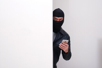 A criminal wearing balaclava holding mobile phone peeking from behind wall