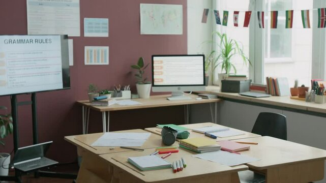 Full no people shot of classroom in English language school, with students copybooks on desks, posters with grammar rules on walls, diverse country flags on windows, computer and multimedia screen