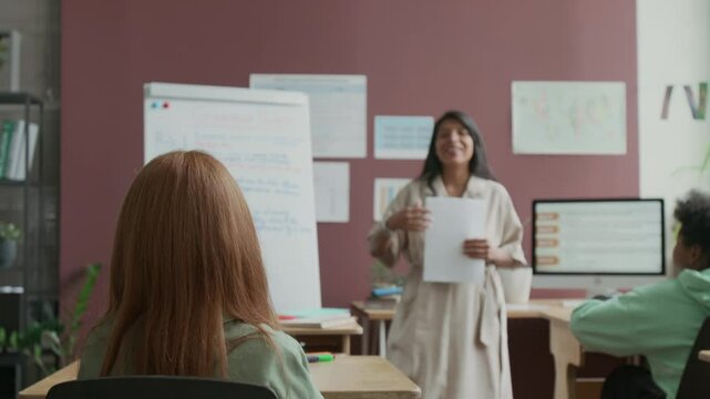 Medium rear shot of young red-haired Caucasian girl raising hand to answer teachers question during English lesson, while sitting in classroom in language school