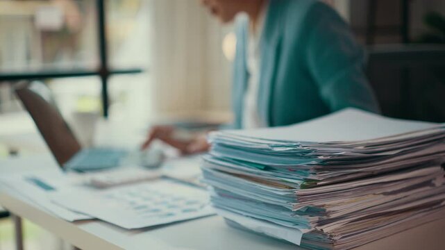 Accountant working diligently at a desk, surrounded by a large stack of invoices. Papers and documents clutter the workspace, reflecting a busy and demanding financial environment