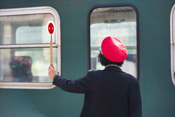 A female traffic controller in a black uniform with a red hat gives a signal to the driver of a green train