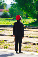 A female traffic controller in a black uniform with a red cap awaits the arrival of a train on the station track
