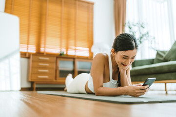 Young Asian woman lying on floor on yoga mat using smartphone at home. Beautiful smiling woman doing exercise relaxing while chatting with trainer online after workout