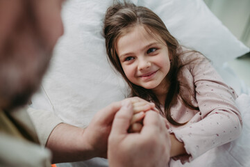 Father beside daughter on hospital bed, holding hands. Sick little girl in hospital.