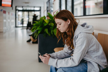 Teenage girl waiting in hospital corridor, sitting on bench and looking at smartphone. Adolescent...
