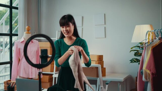 Asian seller woman presents clothes in a bright workshop, showcasing garments in front of a smartphone. The scene highlights a modern approach to online retail and entrepreneurship