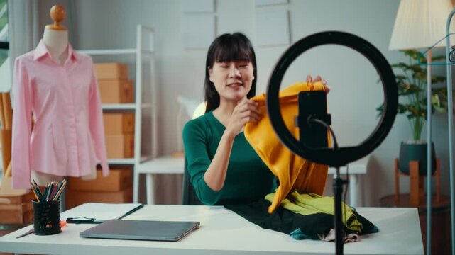 Asian fashion blogger is presenting clothes using her smartphone and a ring light. She is sitting at her desk in her office