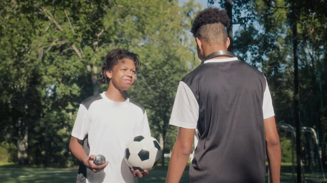 Tilt up back view shot of two African American teen soccer players wearing sports uniform talking while going to field together on green grass in public park - Powered by Adobe