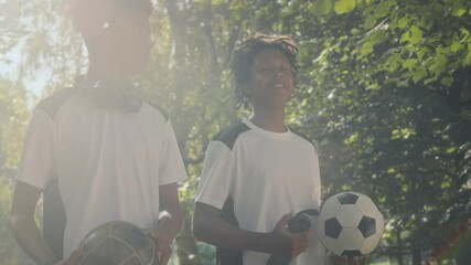 Medium shot of two African American teen male teammates having conversation while going to soccer training together along sunlit green park