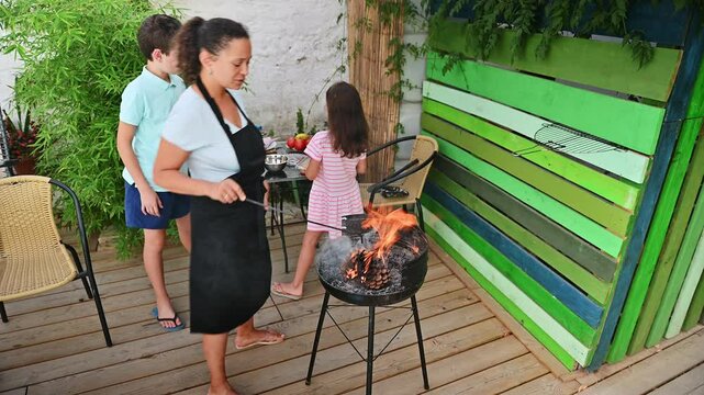 Woman grilling seafood on a barbeque while a child stands nearby in a summer outdoor setting featuring green plants and a cozy wooden deck.