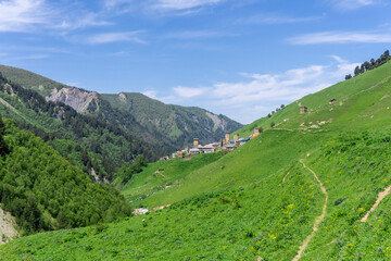 Fototapeta premium Village with towers on a steep slope covered with grass. River at the bottom of the gorge. Bright sky with clouds