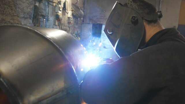 Worker in protective mask welding metal construction at metalworking factory. Welder doing joint between two steel details at industrial production. Man welds parts of the metal detail together