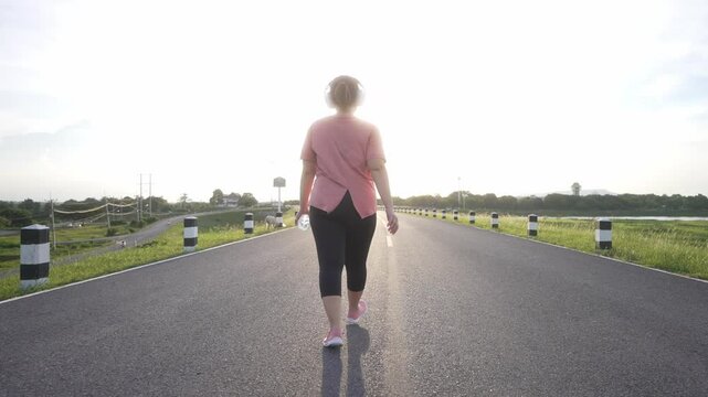 A rear view of a middle-aged, overweight Asian woman walking for exercise on a road in the evening, wearing athletic wear and headphones, holding a water bottle, and embracing a healthy lifestyle