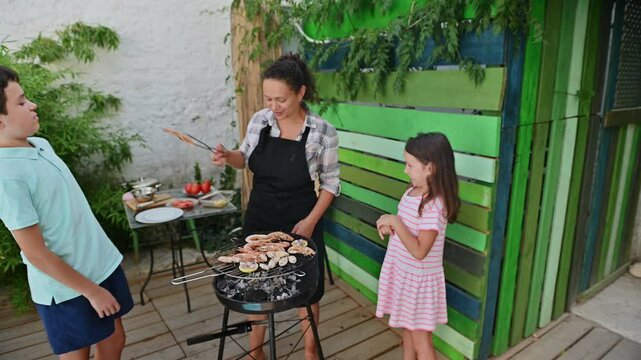 Family enjoying seafood barbeque outdoors with children watching, surrounded by green plants and a colorful painted wall