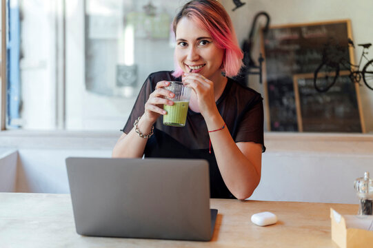 Smiling freelancer with pink hair drinking macha tea and sitting with laptop in cafe