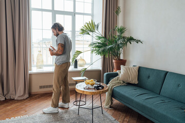 Young man looking at beer bottle standing in front of window at home