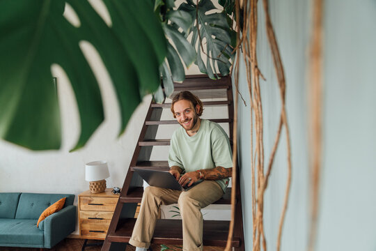 Smiling young freelancer sitting with laptop on staircase at home